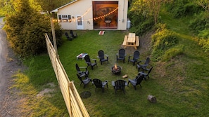 A circular seating arrangement with chairs and a fire pit in the center, surrounded by a wooden deck and a building with string lights.