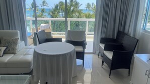 Ein Balkon mit Blick auf das Meer und Palmen, ausgestattet mit einem weißen Sofa, einem runden Tisch und Korbstühlen.