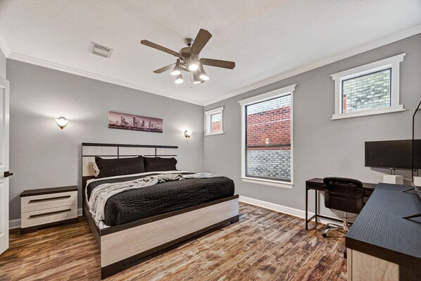 A bedroom with a bed, a desk, a chair, a ceiling fan, and two windows.
