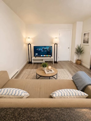 Uma sala de estar com sofá, mesa de centro, televisão, duas luminárias de chão, uma porta, um quadro emoldurado e uma pequena planta.