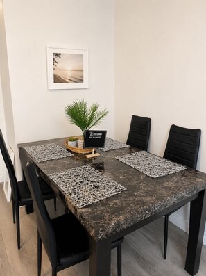 Uma mesa de jantar com tampo de mármore escuro, cadeiras pretas, um jogo americano tecido, uma pequena planta e uma placa de boas-vindas.