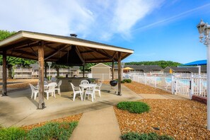 A covered outdoor patio with tables and chairs, a barbecue grill, and a pool area with a white fence. - Waterfront Lake Escape - Pool, Dock, Sunset Views (Osage Beach)