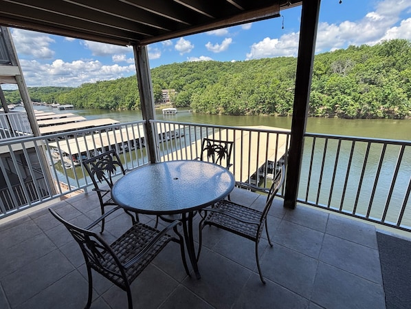A covered outdoor area with a round table and chairs, overlooking a river with boats and a forested hill in the background.