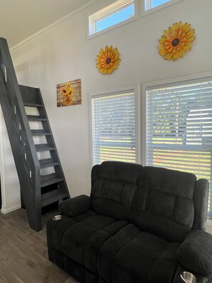 Ein Wohnzimmer mit einem schwarzen Sofa, einer Holzleiter und zwei großen Fenstern mit Jalousien.