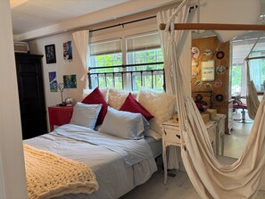 A bedroom with a bed, a bedside table, a mirror, a window with curtains, and a hammock. - The Tipsy Gypsy - Studio Apartment (Brewster)