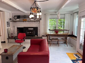A living room with a fireplace, red sofa, dining table, and a view of greenery outside. - The Tipsy Gypsy - Studio Apartment (Brewster)