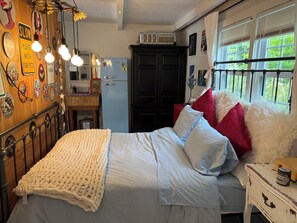 A bedroom with a bed, a dresser, a nightstand, a window with curtains, and a wall with various decorations. - The Tipsy Gypsy - Studio Apartment (Brewster)