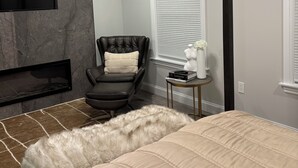 A modern bedroom with a bed, a leather chair, a small table with books, and a TV mounted on a stone wall. - Hamptons Retreat Pool Oasis Modern House (Hampton Bays)