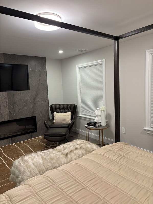 A modern bedroom with a bed, a leather chair, a small table with books, and a TV mounted on a stone wall. - Hamptons Retreat Pool Oasis Modern House (Hampton Bays)