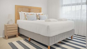 Une chambre à coucher bien rangée, avec un lit, une table de chevet et une fenêtre ornée de rideaux légers.