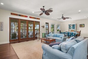 A spacious living room with a large sofa, a coffee table, and a dining area with a table and chairs. There are ceiling fans, a large window with a view, and a sliding glass door leading to an outdoor patio. - Lakefront Getaway on Lake June — Sleeps 12 (Lake Placid)
