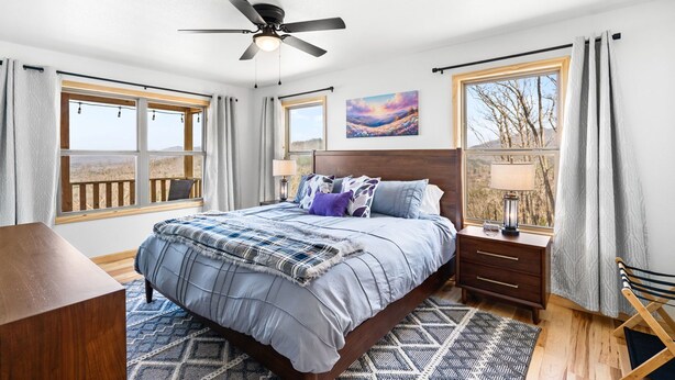A bedroom with a bed, a ceiling fan, a nightstand, a lamp, a window with curtains, and a painting on the wall. - NEW CABIN-Epic Sunsets! Panoramic Views of Blue Ridge and Smokey Mountains! (Murphy)