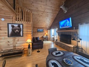 Interior de una cabaña con revestimiento de madera, televisor, chimenea, sofá y mesa de comedor.
