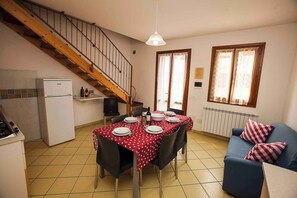 A dining area with a table set for four, a kitchen with a refrigerator, a blue sofa, and a wooden staircase. - Holiday apartment with pool (Rosolina Mare)
