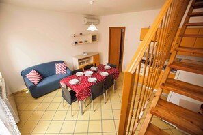 A dining area with a table set for four, a blue sofa with red and white pillows, a wooden staircase, and a kitchen in the background. - Holiday apartment with pool (Rosolina Mare)