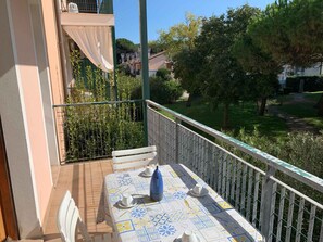 A balcony with a table set for two, a chair, and a view of a garden and buildings. - Holiday apartment with air conditioning (Rosolina Mare)