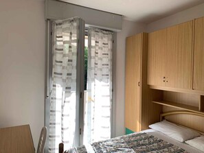 A bedroom with a bed, wooden wardrobe, and a sliding glass door with patterned curtains. - Holiday apartment in a residence with pool (Rosolina Mare)