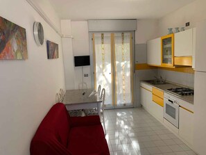 A compact kitchen with a dining area, a red sofa, and a TV mounted on the wall. - Holiday apartment in a residence with pool (Rosolina Mare)