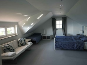 Ein Schlafzimmer mit schrägverdeckter Decke, zwei Betten, einem Schlafsofa, einem Nachttisch und einem fenster mit Vorhängen.