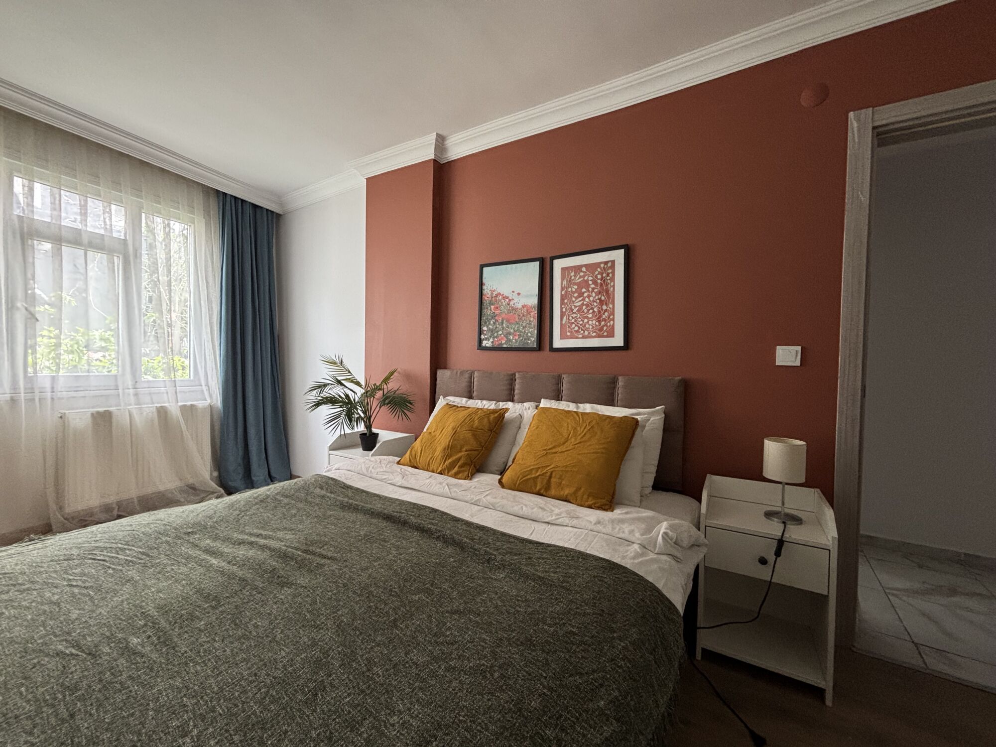 Quarto com cama, mesa de cabeceira, luminária, janela com cortinas e duas imagens emolduradas na parede.