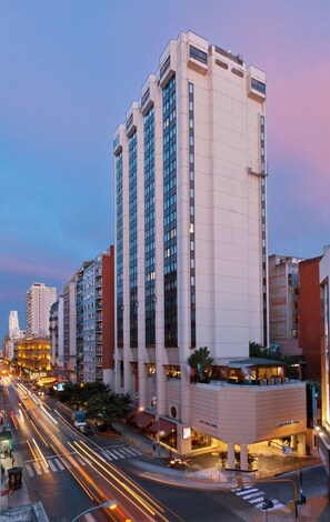 Un hotel moderno de gran altura, con varios pisos, una entrada destacada y una calle bien iluminada en primer plano.