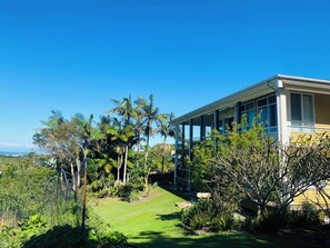 A modern house with a large glass window, surrounded by lush greenery and palm trees.