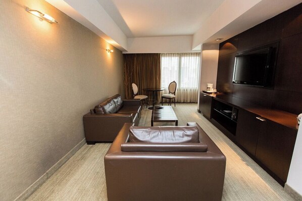 Una habitación de hotel moderna con sofá, una pequeña mesa de comedor, dos sillas, televisor y minibar.