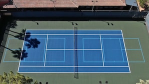 Una cancha de tenis al aire libre con red, rodeada de árboles y un edificio con tejado de tejas.