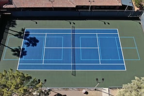 An outdoor tennis court with a net, surrounded by trees and a building with a tiled roof.