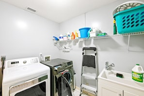 Een wasserette met een wasmachine, droger, planken met schoonmaakmiddelen, een opvouwtafel en een spoelbak.