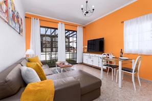 Ein Wohnzimmer mit einer Couch, einem Esstisch mit Stühlen, einem Fernseher und einem großen Fenster mit Vorhängen.