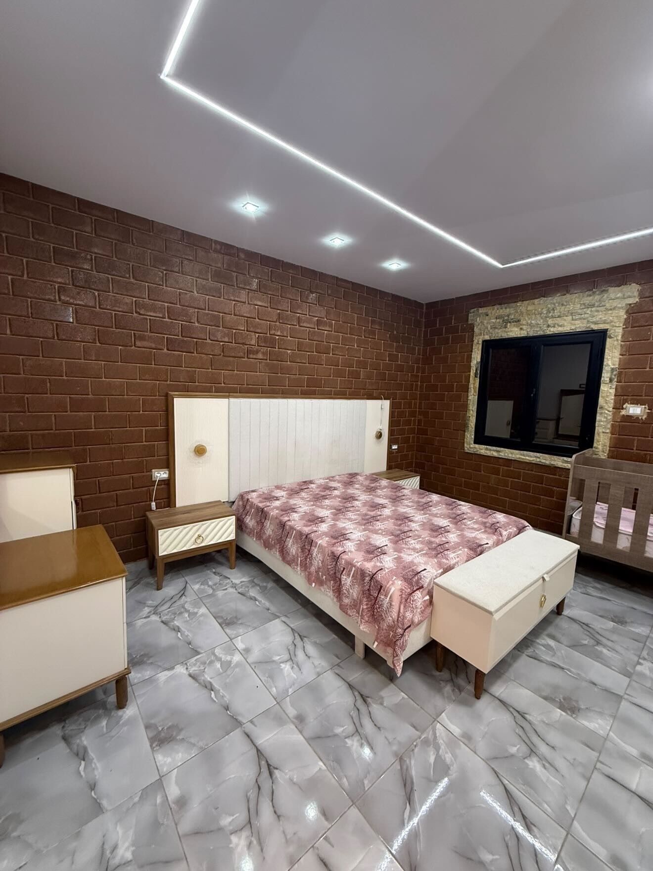 Un dormitorio con pared de ladrillo, una cama con colcha estampada, una mesita de noche, un aparador y una ventana con vistas.