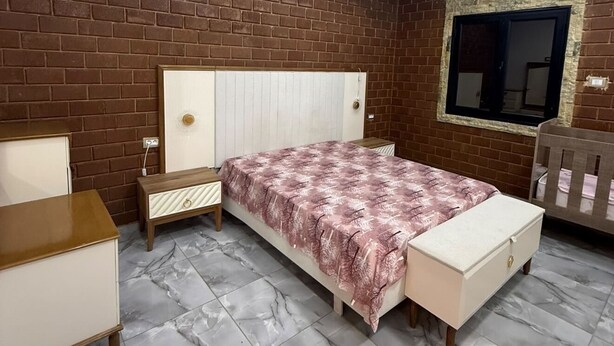 Um quarto com parede de tijolos, uma cama com colcha estampada, uma mesa de cabeceira, cômoda e uma janela com vista.