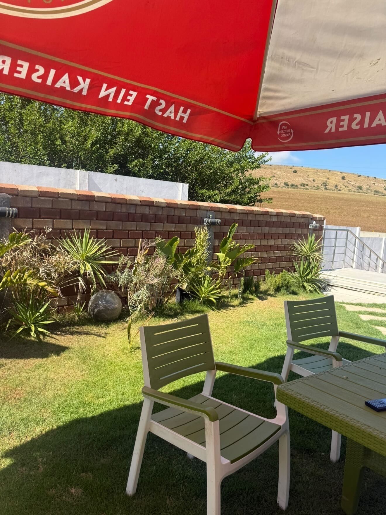 A patio area with two green plastic chairs, a table, and a red umbrella.