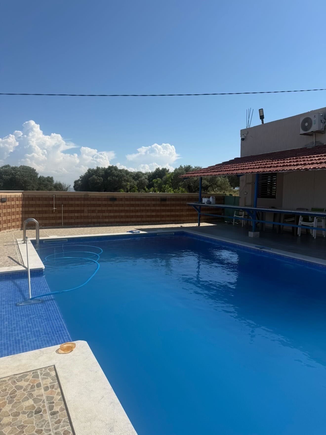 A swimming pool with a tiled deck, a brick wall, and a building with a red-tiled roof.
