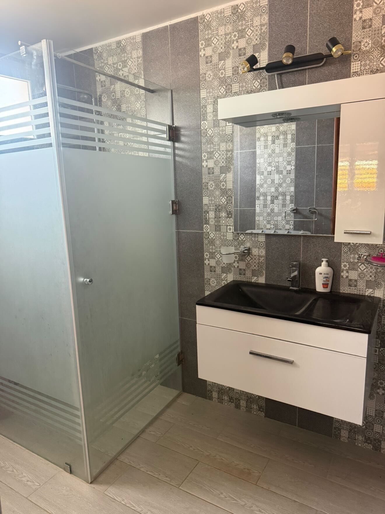 A modern bathroom with a glass shower enclosure, a white vanity with a black countertop, and patterned tiles on the walls.