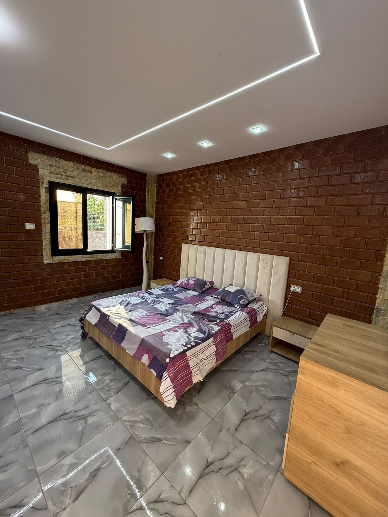 Une chambre avec un mur en briques, un lit recouvert d’une couvre-lit à motifs, une table de chevet et une lampe.