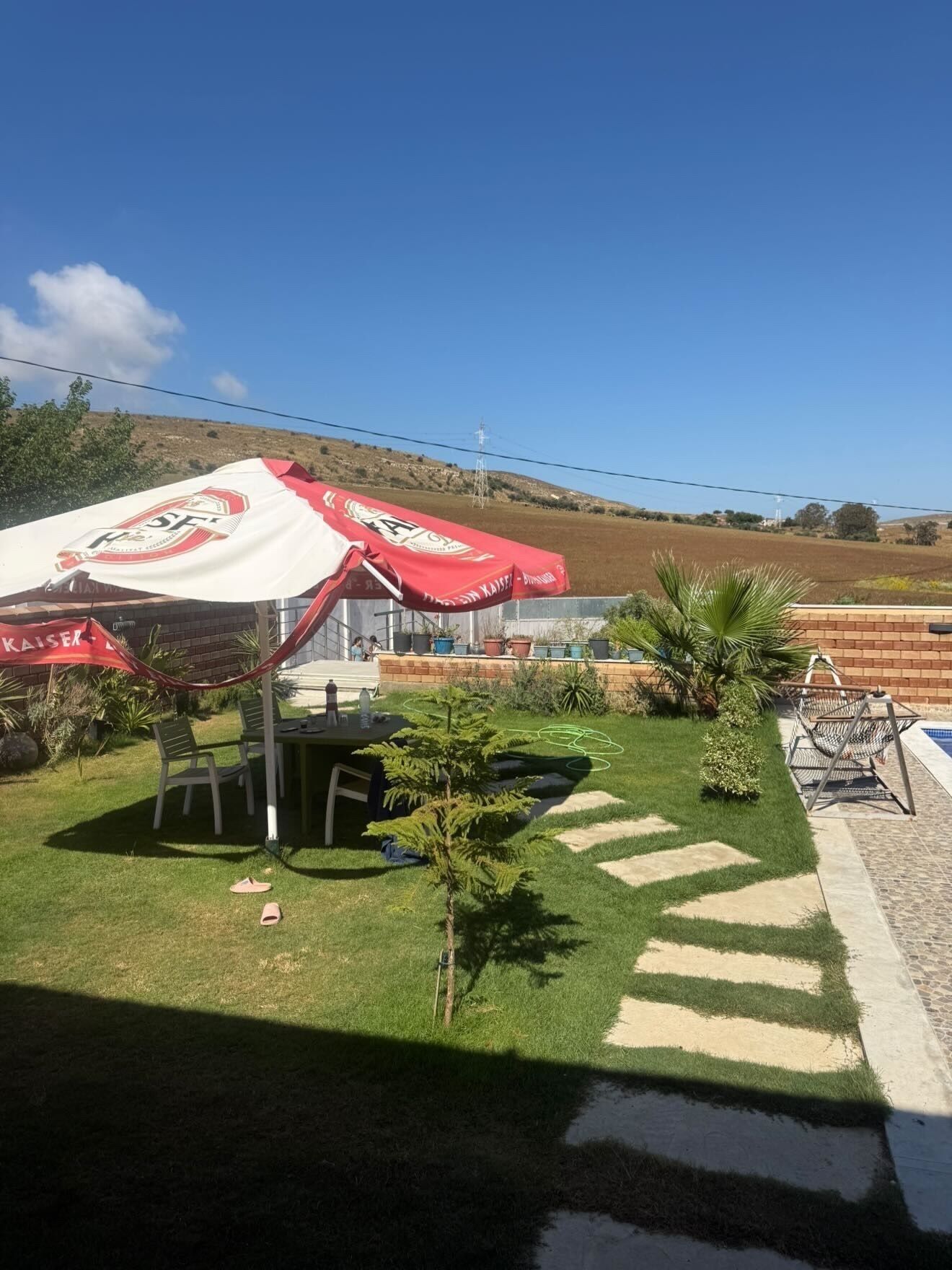 A patio area with a table and chairs, a small garden with plants, and a view of a hill in the background.