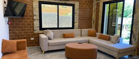 A modern living room with a sectional sofa, a round ottoman, and a flat-screen TV mounted on a brick wall.