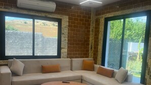 Uma sala de estar moderna com um sofá seccional, um banquinho redondo e grandes janelas com vista para o exterior.