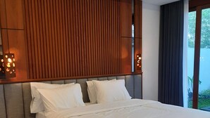 A modern bedroom with a large bed, wooden headboard, bedside lamps, and a view of greenery outside. - JJ Villa 7 Ocean Blue (Badung)