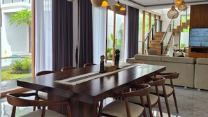 A modern dining area with a wooden table and chairs, a staircase, and a TV mounted on the wall. - JJ Villa 7 Ocean Blue (Badung)