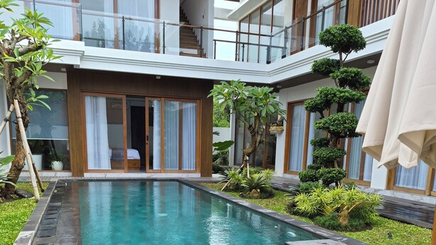 A modern outdoor pool area with a wooden deck, lounge chairs, and a two-story building with large windows. - JJ Villa 7 Ocean Blue (Badung)