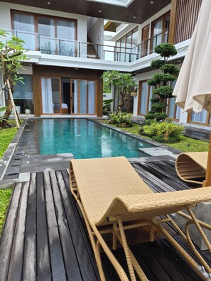 Un área de piscina moderna al aire libre con cubierta de madera, sillones reclinables y un edificio de dos pisos con amplias ventanas.