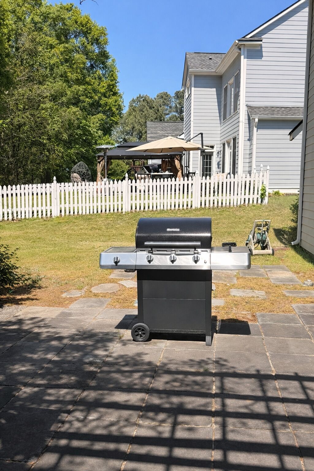 A barbecue grill in a backyard with a white picket fence, a house, and a patio area.