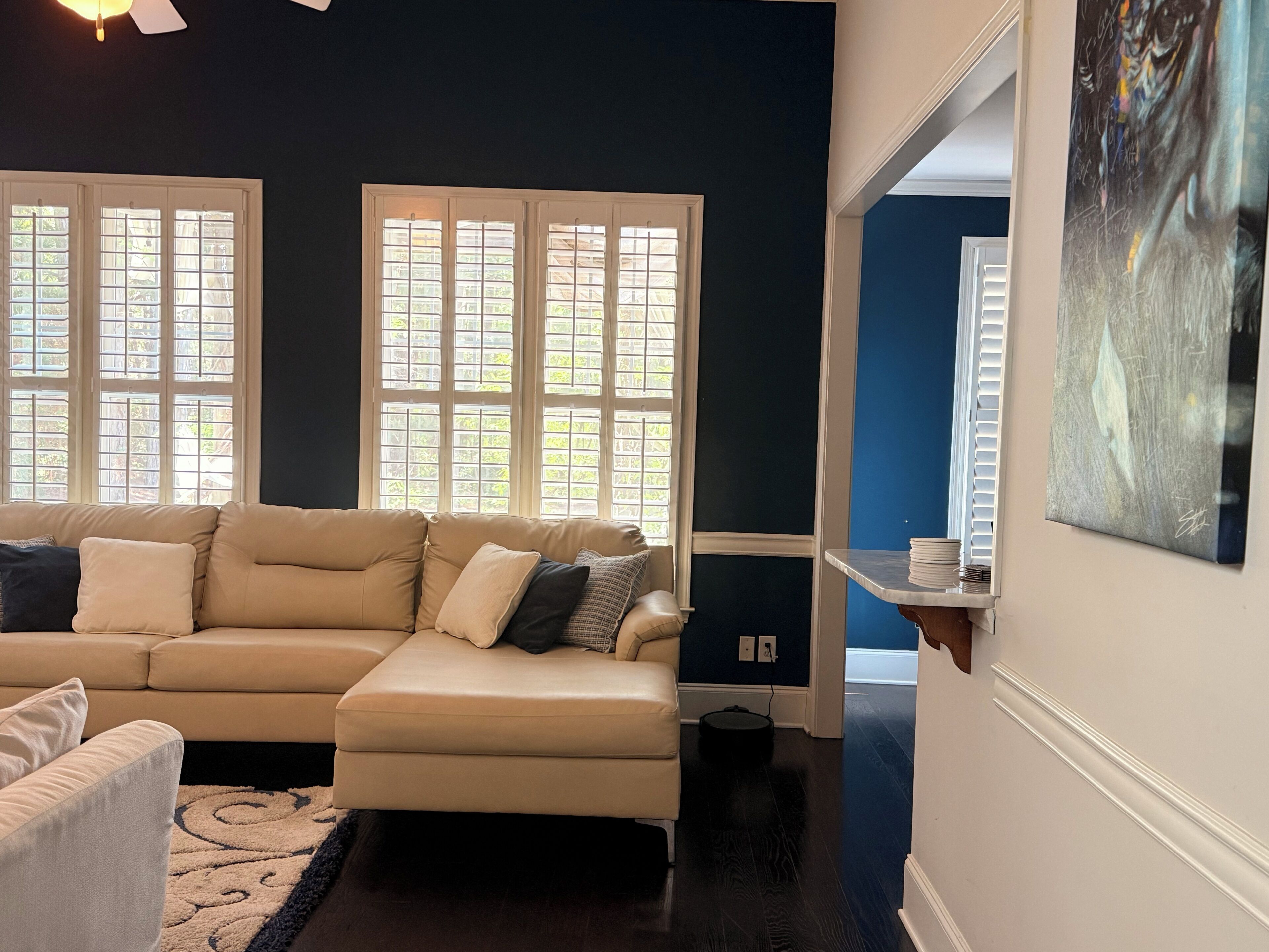 A living room with a beige sectional sofa, two windows with blinds, a dark floor, and a painting on the wall.