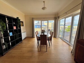 En rummelig stue med spisebord, stole, en bogreol og store vinduer med udsigt over byen.