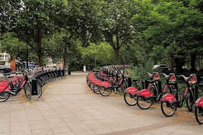 Eine Reihe roter Santander Fahrräder, abgestellt entlang eines von Bäumen gesäumten Bürgersteigs.