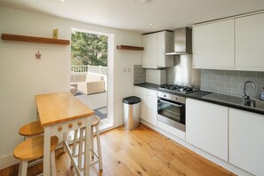Una cocina moderna con armarios blancos, una estufa a gas, horno y lavaplatos. Hay una mesa de madera y sillas, además de un cubo de basura.