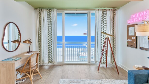 Ein Zimmer mit Schreibtisch, Stuhl, Stativ, einer Neonreklame und einem Balkon mit Meerblick.